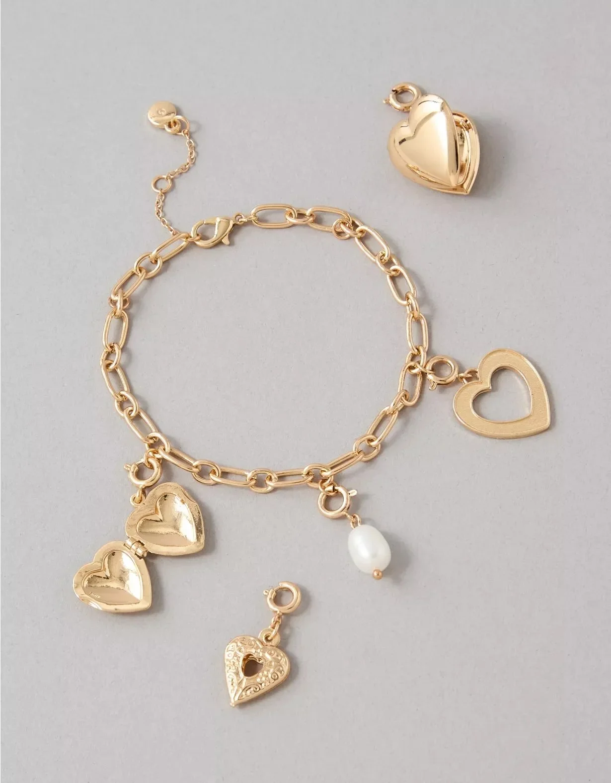 Gold bracelet and matching gold heart-shaped and open-heart charms, along with a pearl and a silver heart-shaped jewelry piece, on a light gray background.