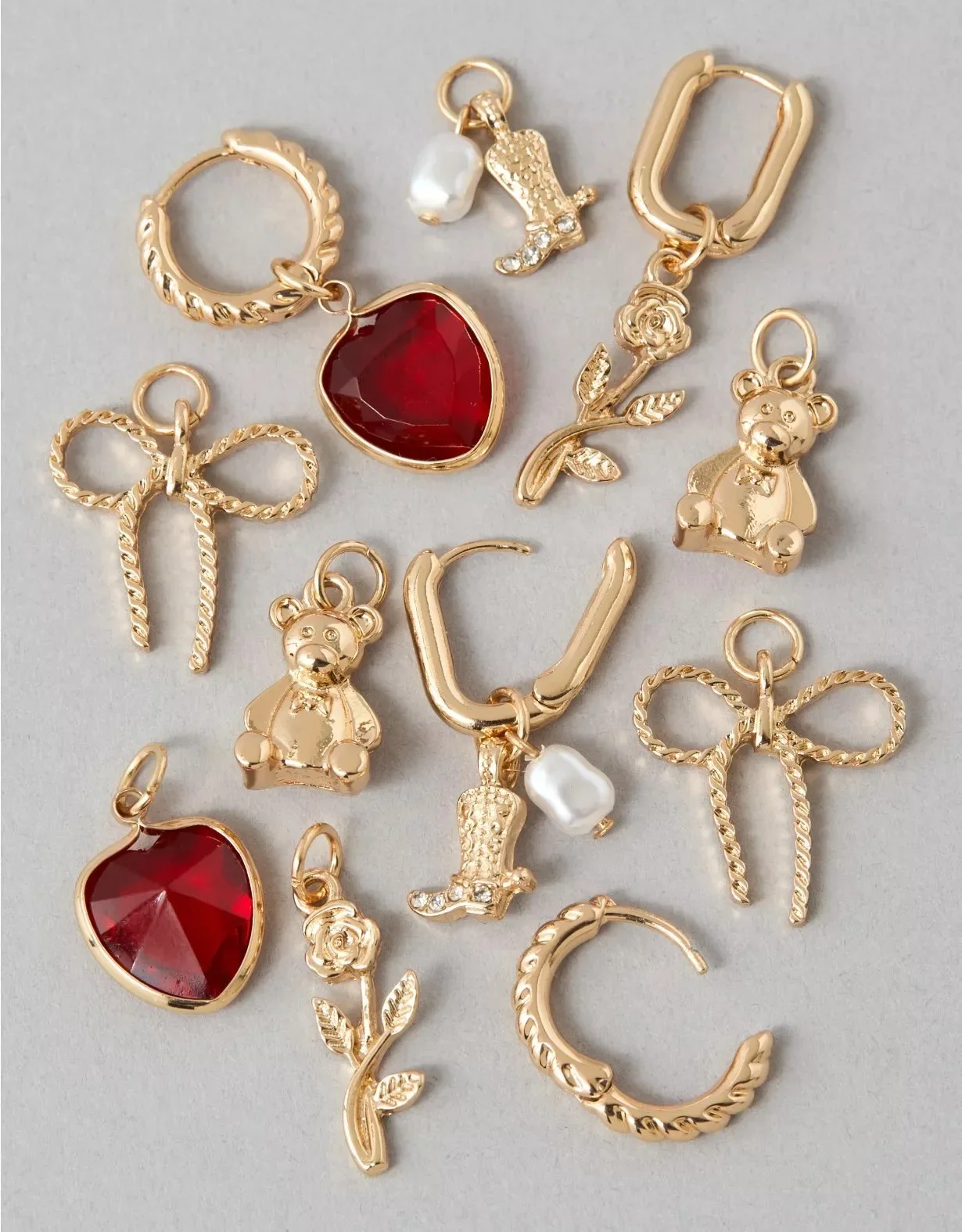 Gold jewelry charms including a red heart, teddy bear, rose with leaves, bow, horseshoe, crescent moon, and a pearl, arranged on a light gray background.