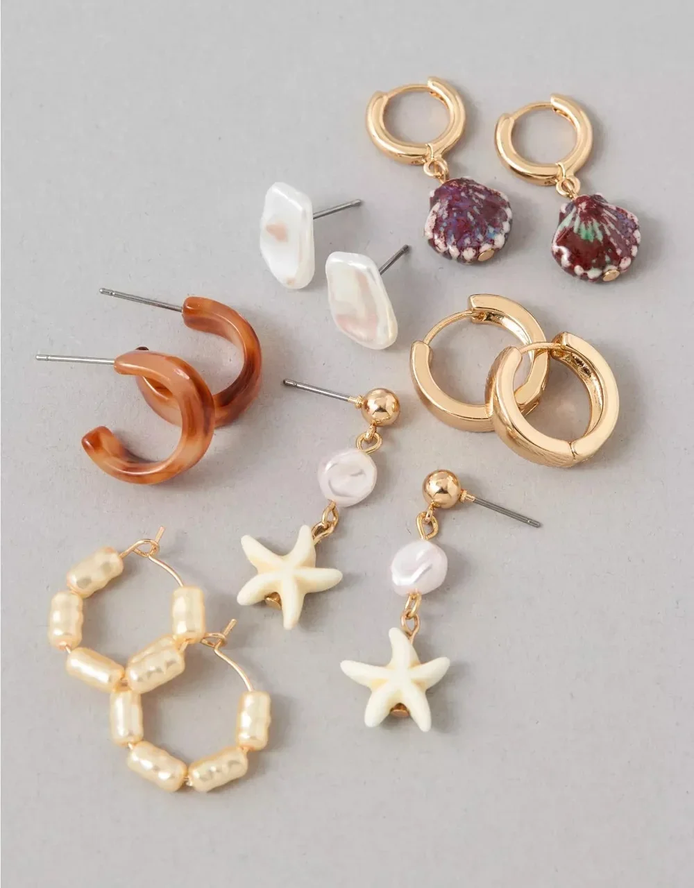 Assorted earrings including gold hoops with purple floral charms, pearl drop earrings with starfish pendants, pearl cluster hoops, tortoise shell hoops, and white pearl studs, arranged on a light gray background.