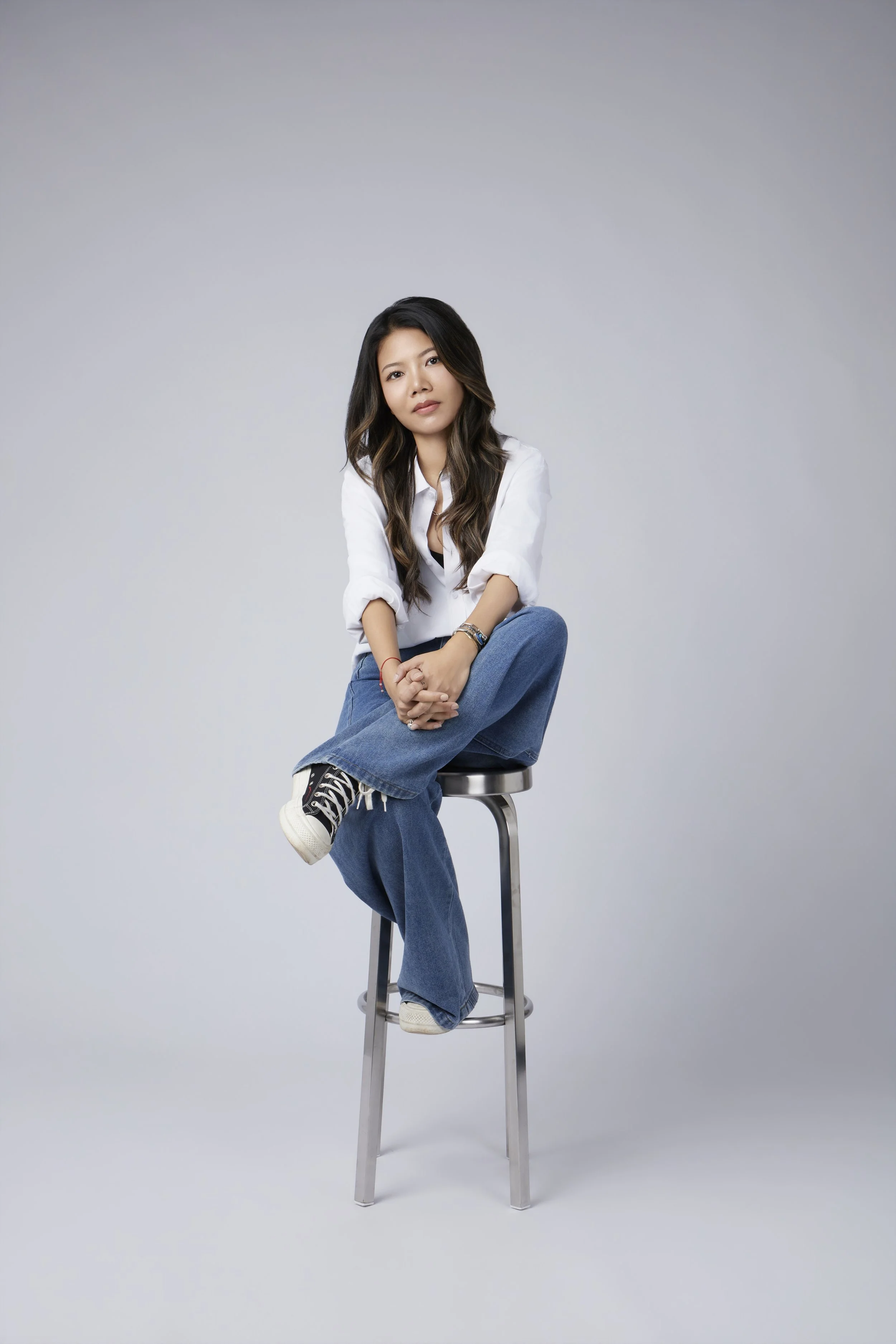 A woman with dark wavy hair sitting on a metal stool against a gray background, wearing a white shirt, blue jeans, and sneakers.