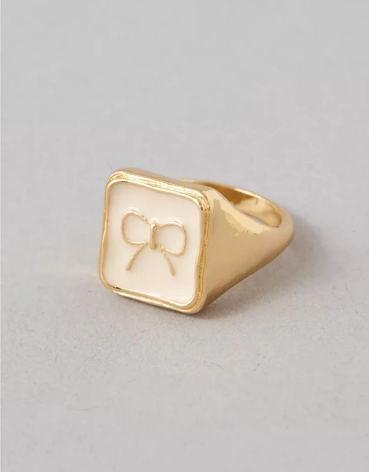 A gold ring with a rectangular face featuring a small engraved bow design on a plain gray background.