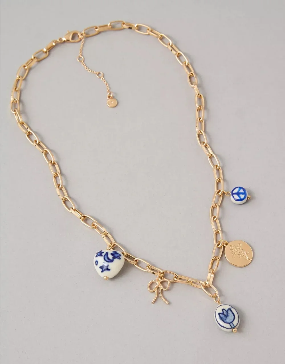 Gold chain necklace with blue and white ceramic charms, gold bow and round gold pendant, on a light grey background.