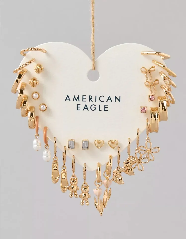 Gold earrings and jewelry charms displayed on a white heart-shaped card with 'American Eagle' written on it, suspended by a string.