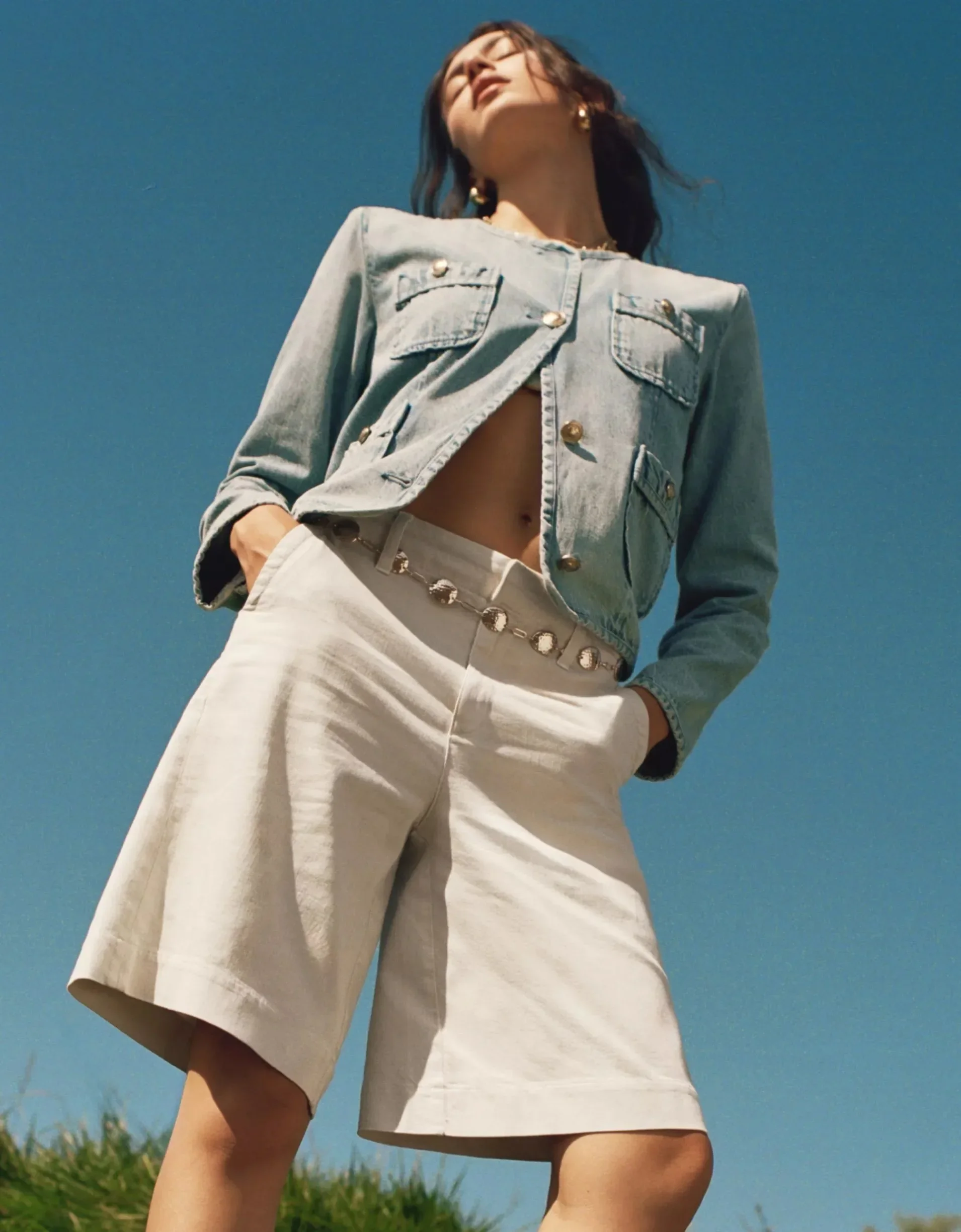 A woman wearing a denim jacket and cream-colored pants stands outdoors against a bright blue sky, with green trees visible at the bottom of the image.