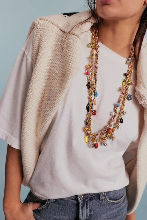 A woman wearing a white T-shirt with a multicolored beaded necklace, a beige knitted vest draped over her shoulders, and blue jeans. Only part of her face is visible.