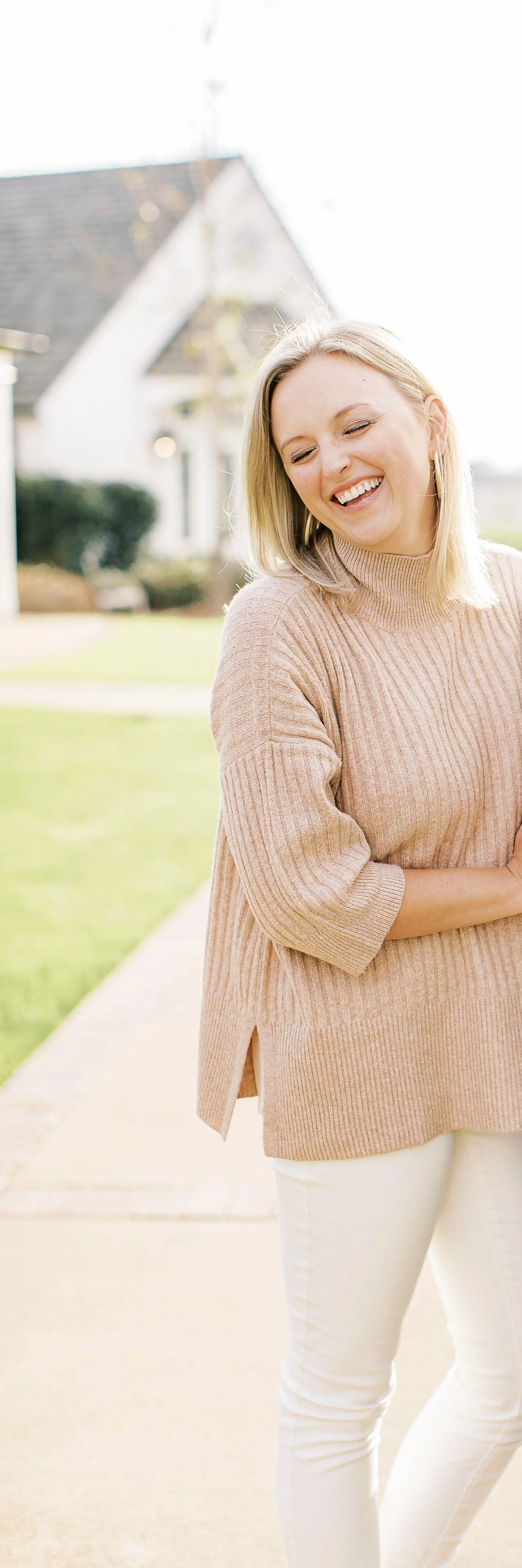 A woman with blonde hair smiling and standing outdoors in a sunny neighborhood, wearing a beige turtleneck sweater and white pants.