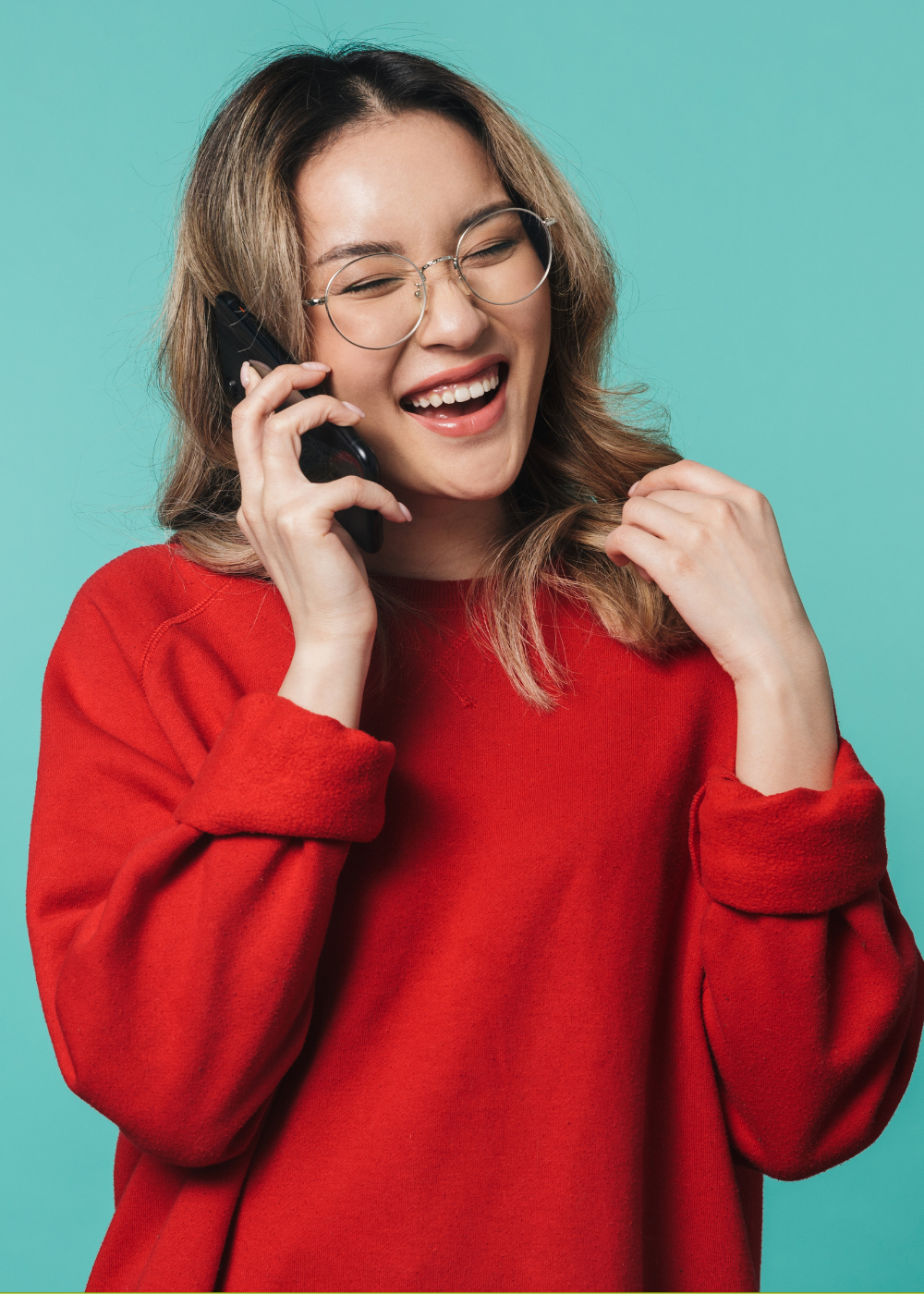 A young woman with wavy brown hair, glasses, and a red sweater, smiling and talking on a phone.