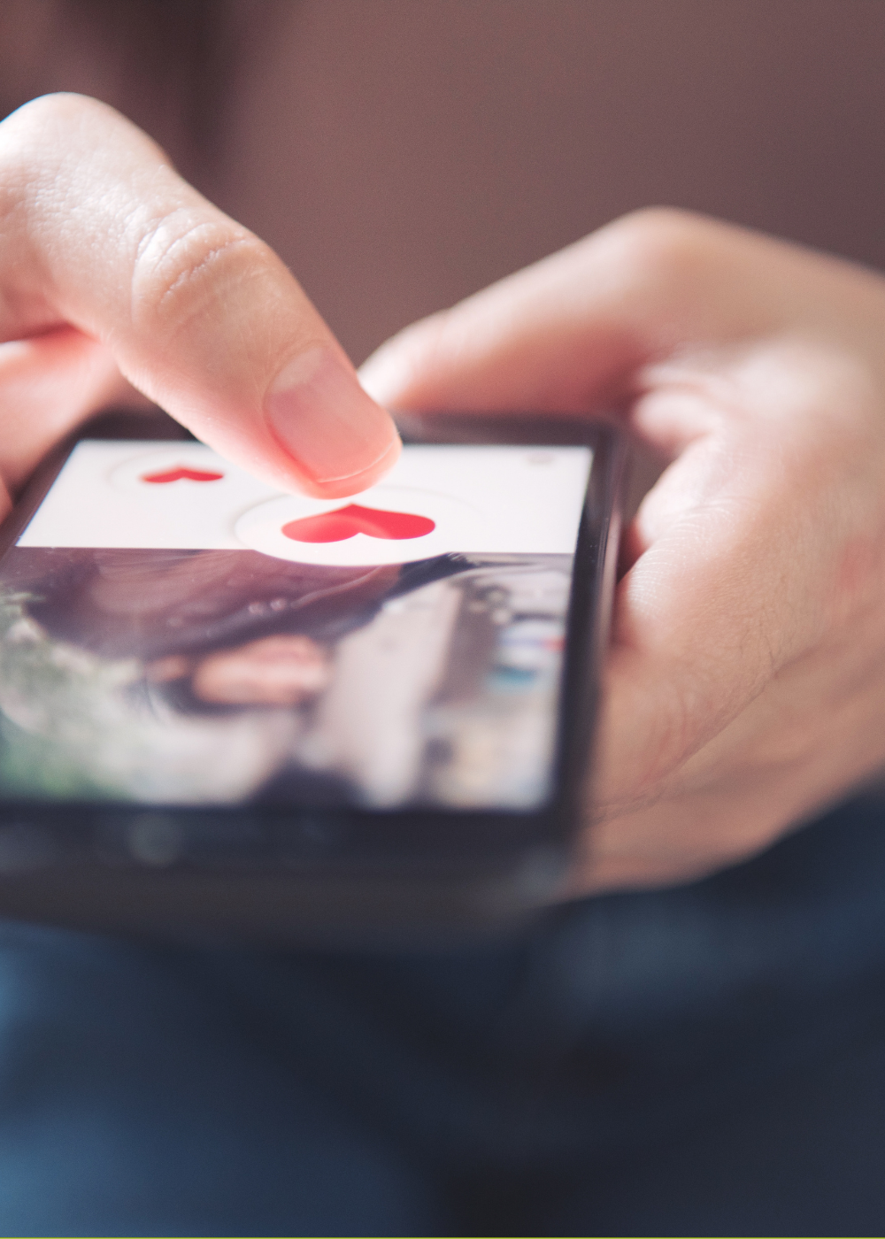 Close-up of a person holding a smartphone, with a finger tapping on the screen displaying red hearts and a blurred background.