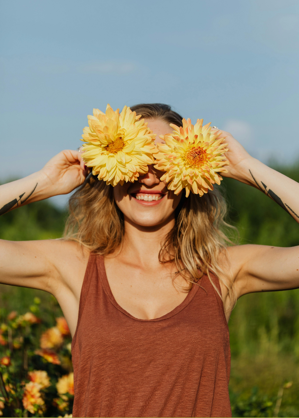 A woman smiling outdoors holds large yellow flowers over her eyes. She wears a sleeveless brown top, has wavy blonde hair, and tattoos on her arms. The background is a bright, sunny sky with blurred greenery.