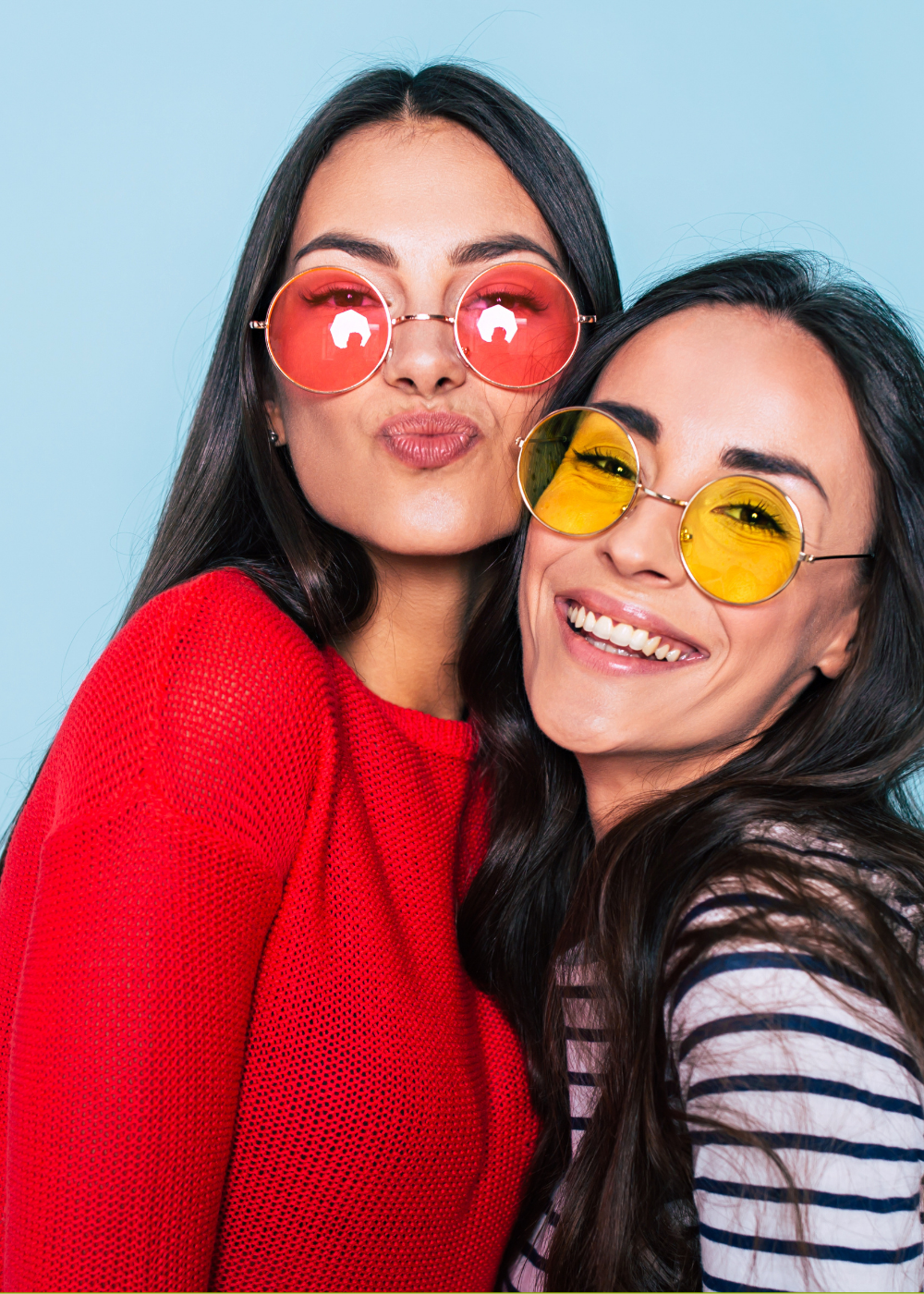 Two women wearing colorful sunglasses and smiling, standing close together against a light blue background.