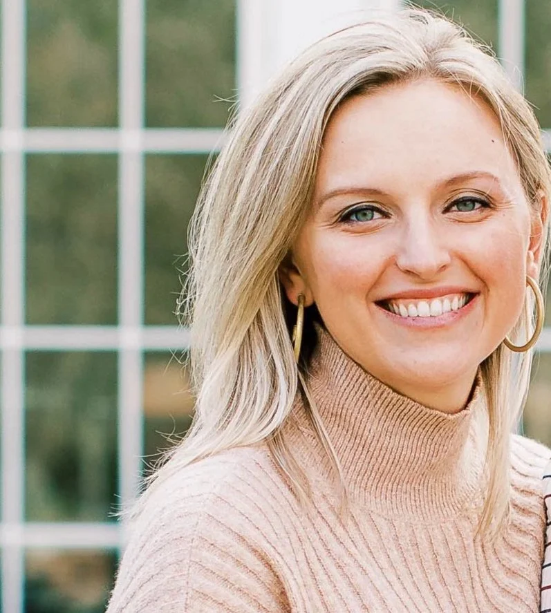 A smiling woman with blonde hair wearing a beige turtleneck sweater and gold hoop earrings, standing outdoors in front of a large window with a grid pattern.