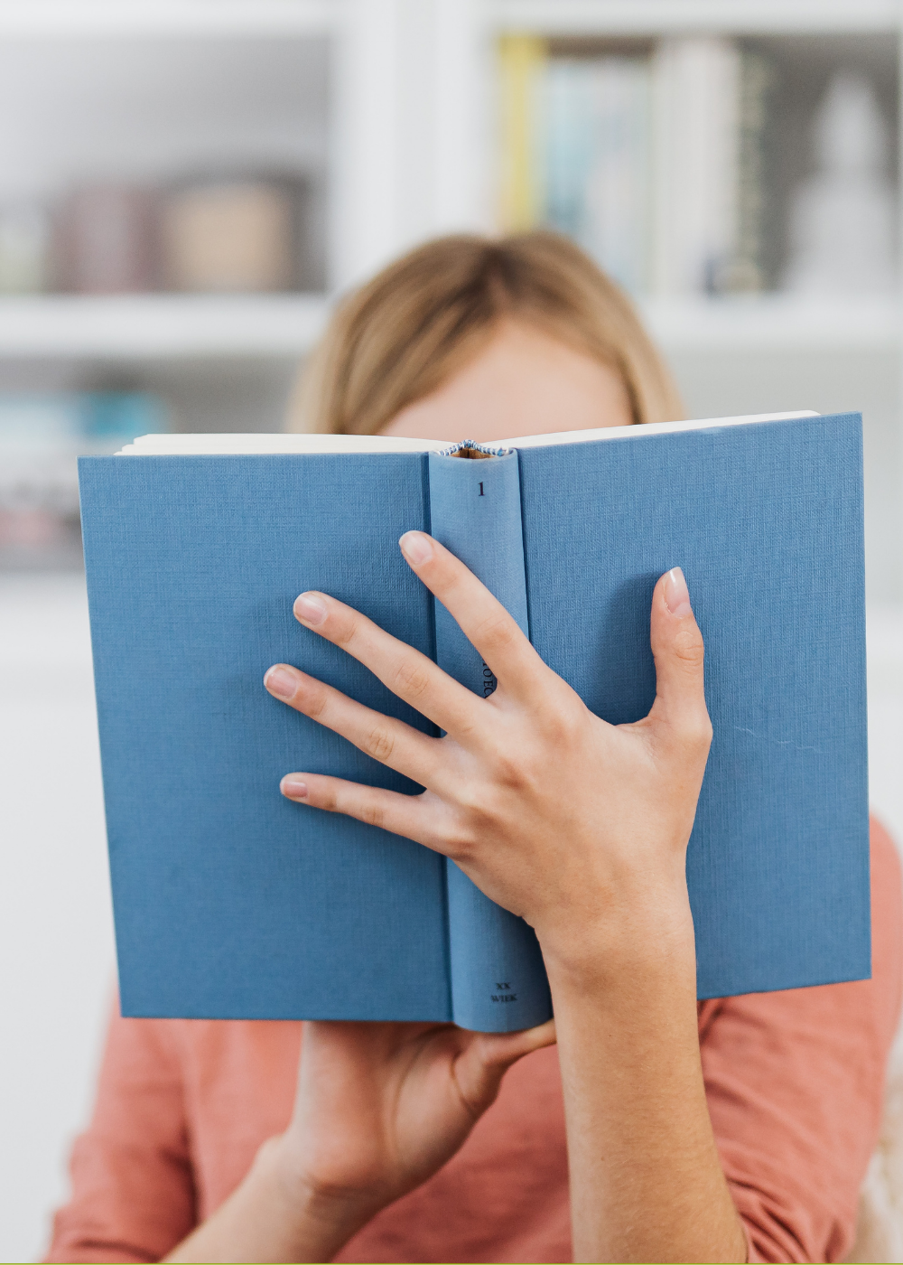 Person holding a blue book in front of their face, with only their hands and part of their head visible in the background.
