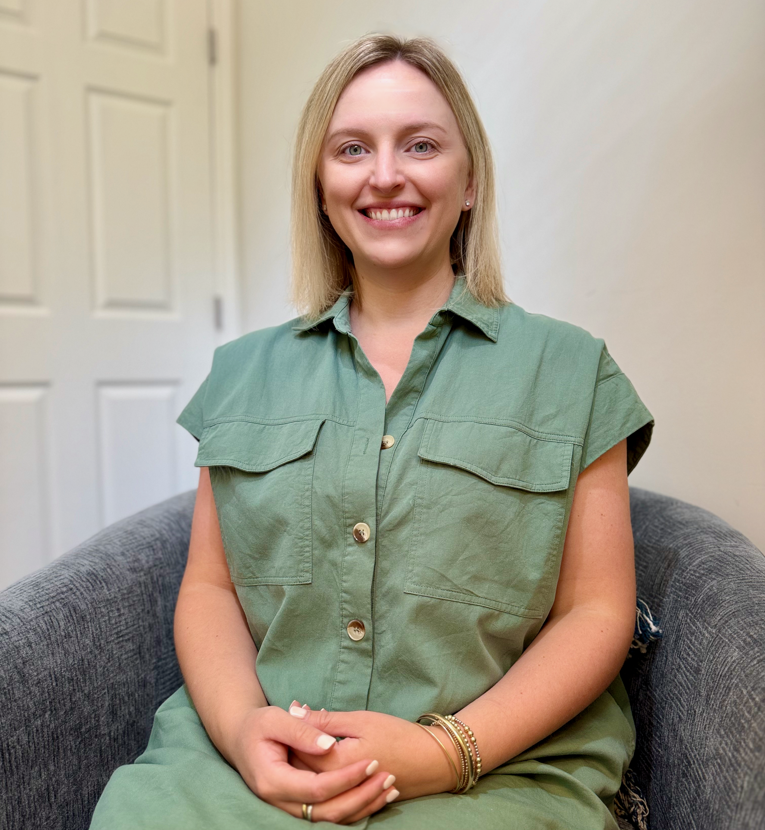 A woman with blonde hair wearing a green button-up dress, sitting on a gray chair, smiling at the camera in a room with white walls.