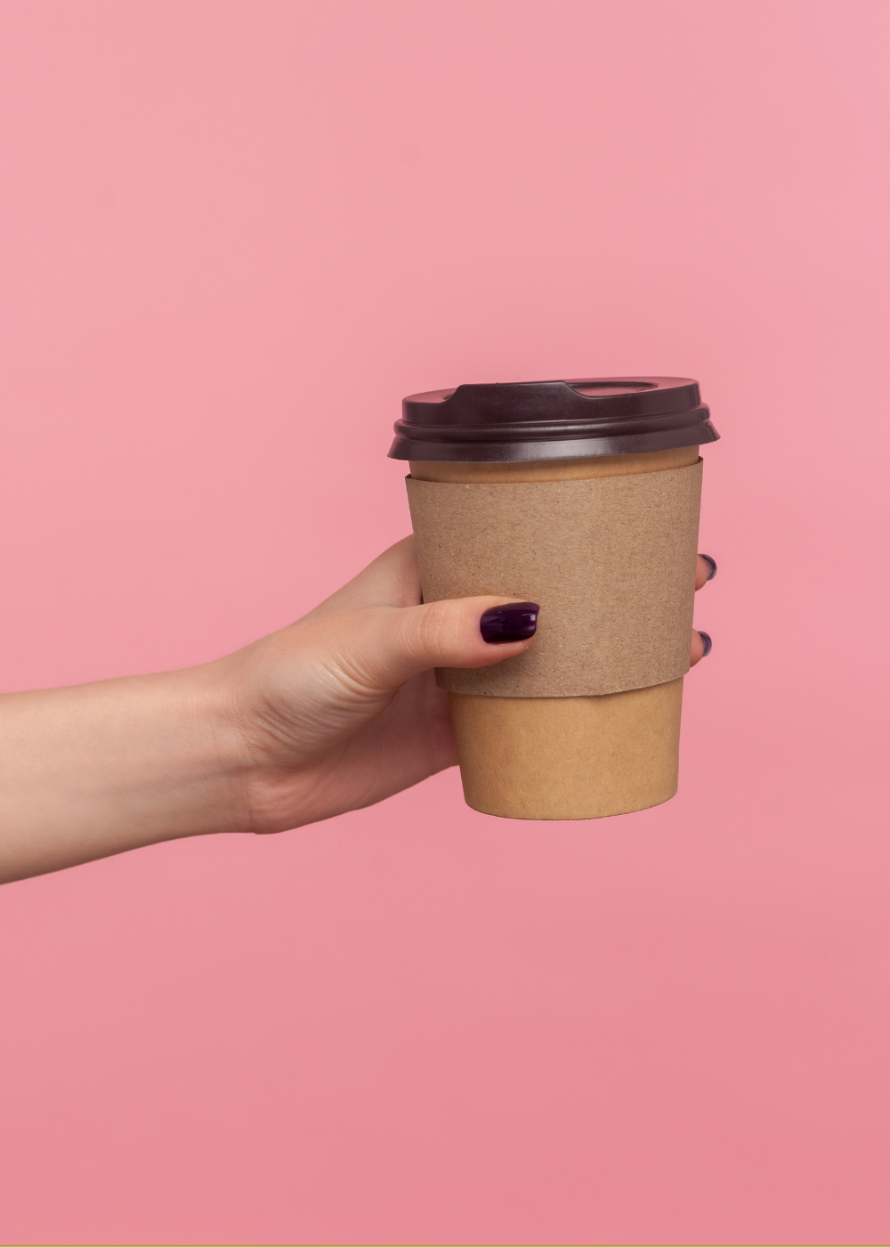 Person holding a disposable coffee cup with a brown cardboard sleeve and dark plastic lid against a pink background.