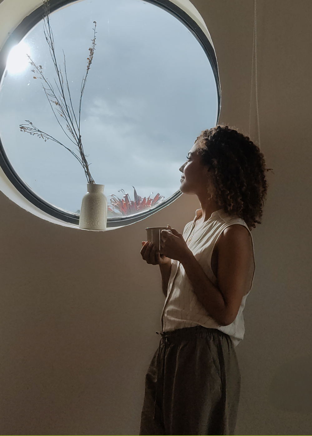 A woman with curly hair holding a cup, standing indoors near a round window with a vase and dried branches on the windowsill, looking outside at a cloudy sky.