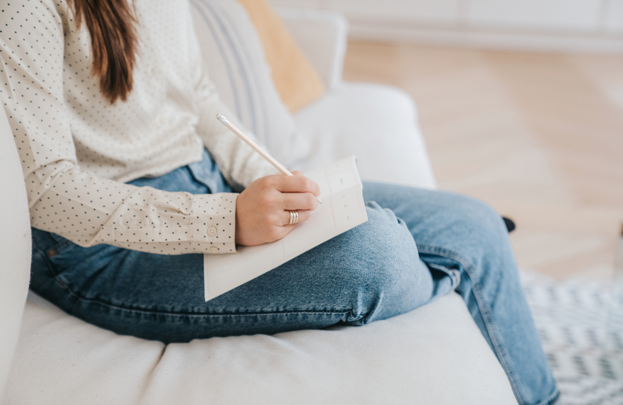 Person sitting on a white sofa, writing in a planner with a pencil, wearing a white polka dot shirt and blue jeans.