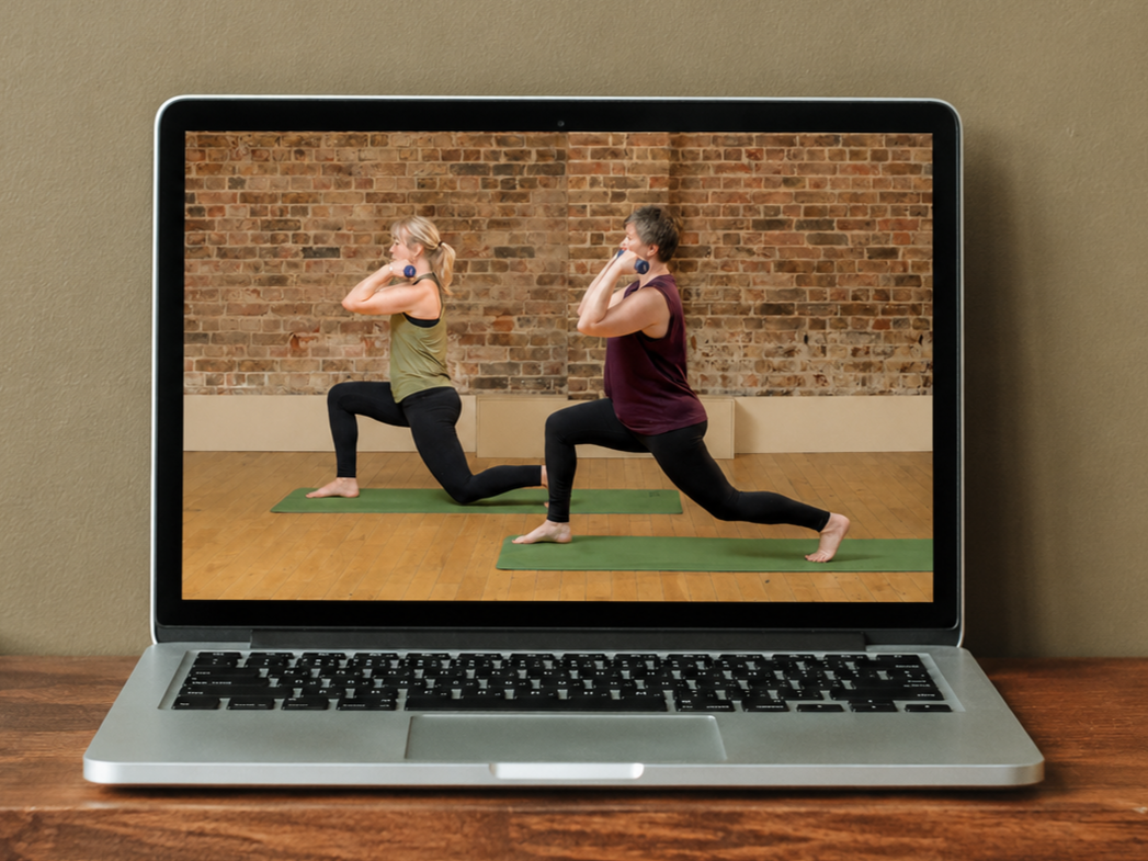 Two women practicing yoga in a studio with brick wall background, using green mats, in a lunge pose holding dumbbells.