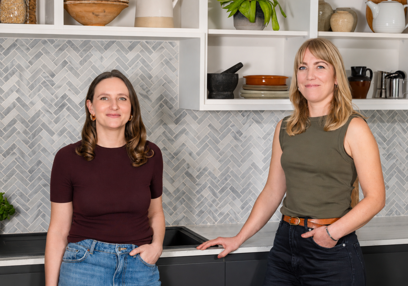 Two women standing in a kitchen with open shelves, various dishes, bowls, and plants on the shelves, a grey and white herringbone tile backsplash, and countertop.