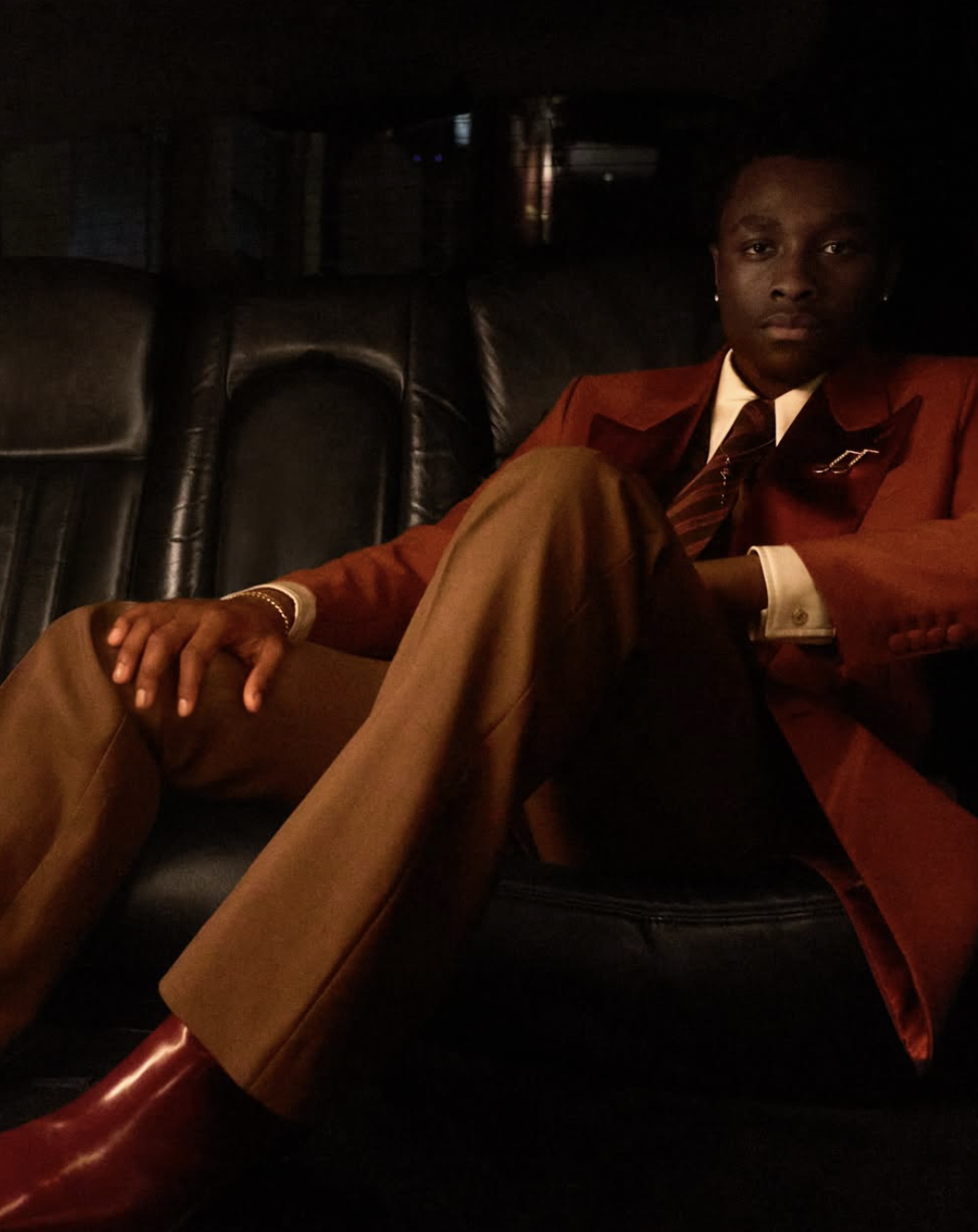 A young man dressed in a brown suit and tie, sitting on a black leather couch in a dimly lit room.