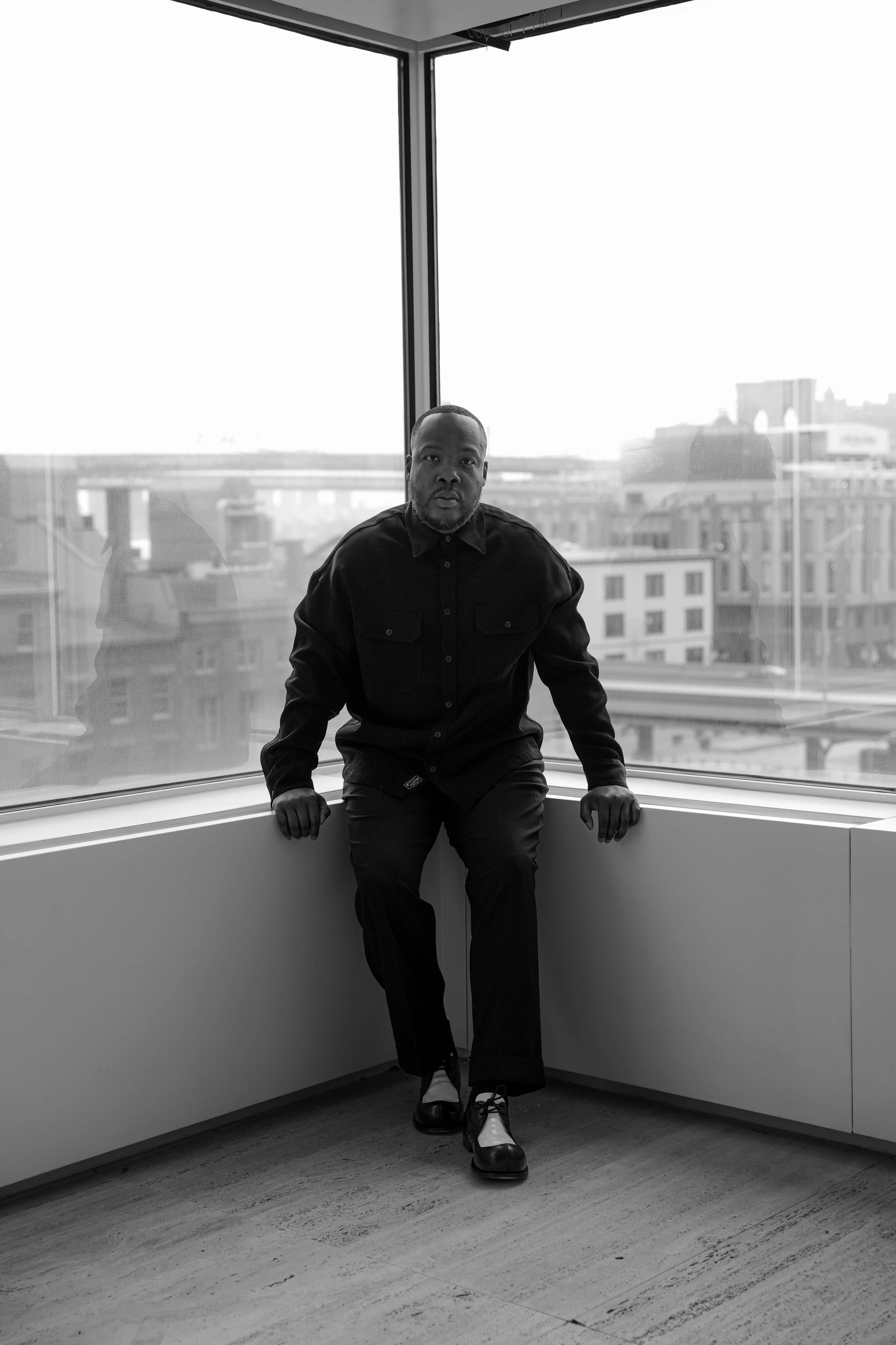 A man dressed in dark clothing and two-tone shoes is sitting on a windowsill inside a modern building, with city buildings visible through large windows in the background.