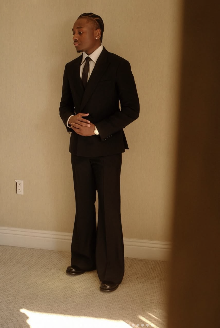 A man in a black suit with a white dress shirt and black tie, standing against a beige wall, with short braided hair, earrings, and a watch, on a beige carpeted floor.
