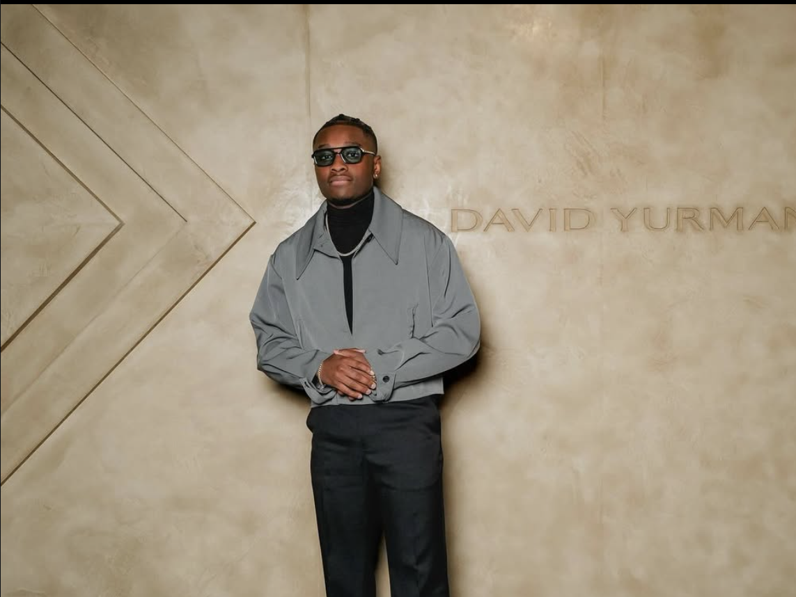 A young man with dark skin, sunglasses, and braided hair stands against a beige wall with the names 'David Yurman' on it. He is wearing a gray jacket, black turtleneck, black pants, and accessorizes with jewelry.