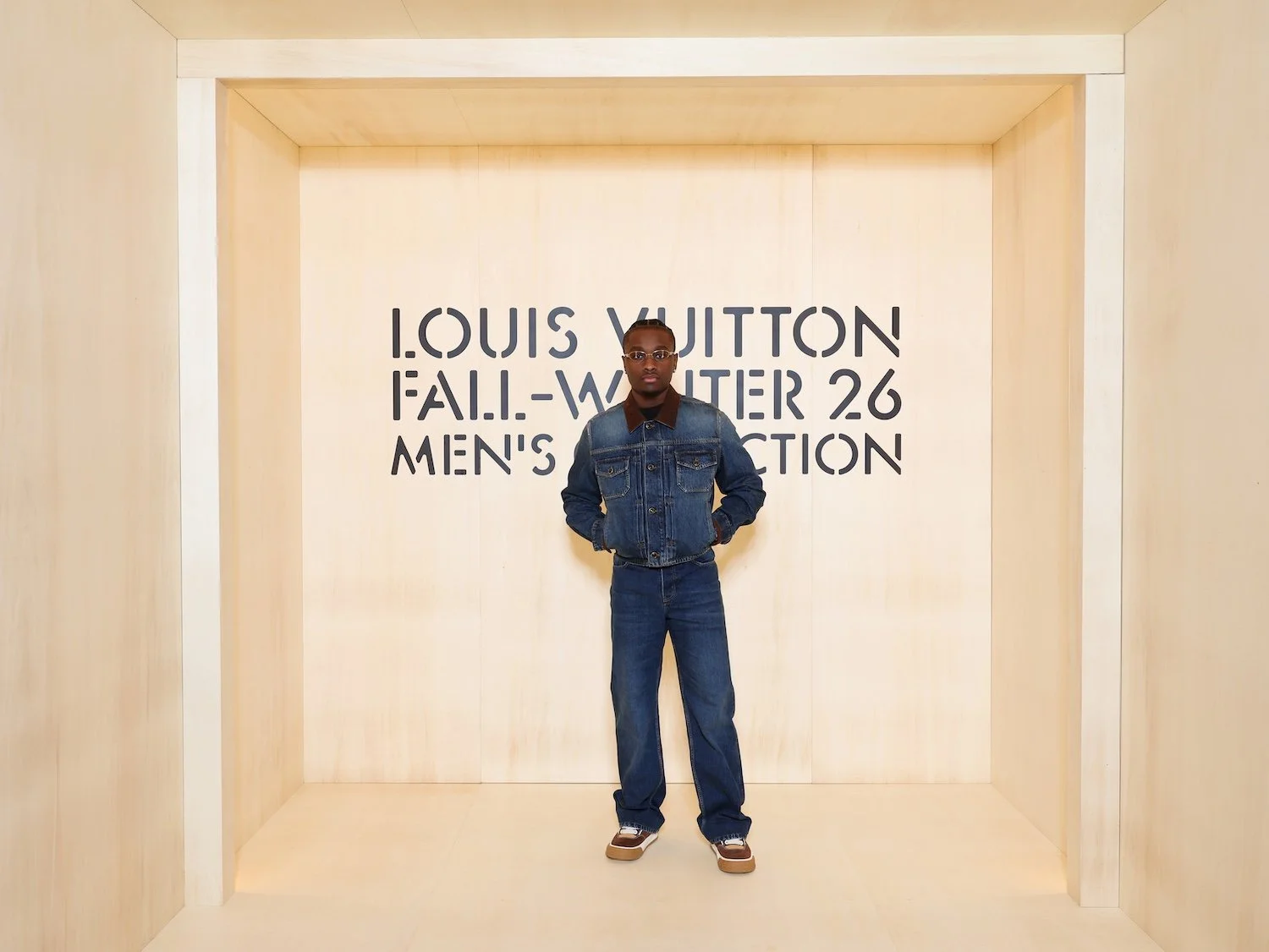 A person standing in front of a sign that says Louis Vuitton Fall-Winter 26 Men's Collection, wearing a denim jacket and pants with a backpack, in a light-colored room.
