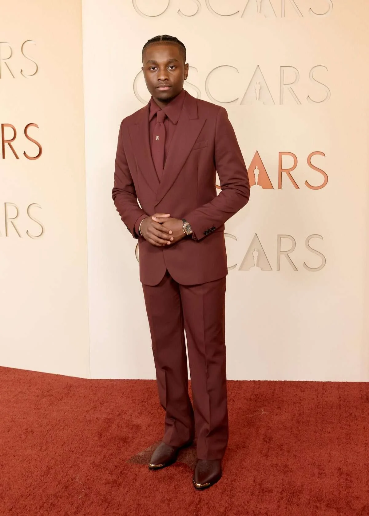 A young man in a maroon suit standing with hands clasped in front of him, on a red carpet with a white background featuring the words "Oscars" in different fonts and colors.