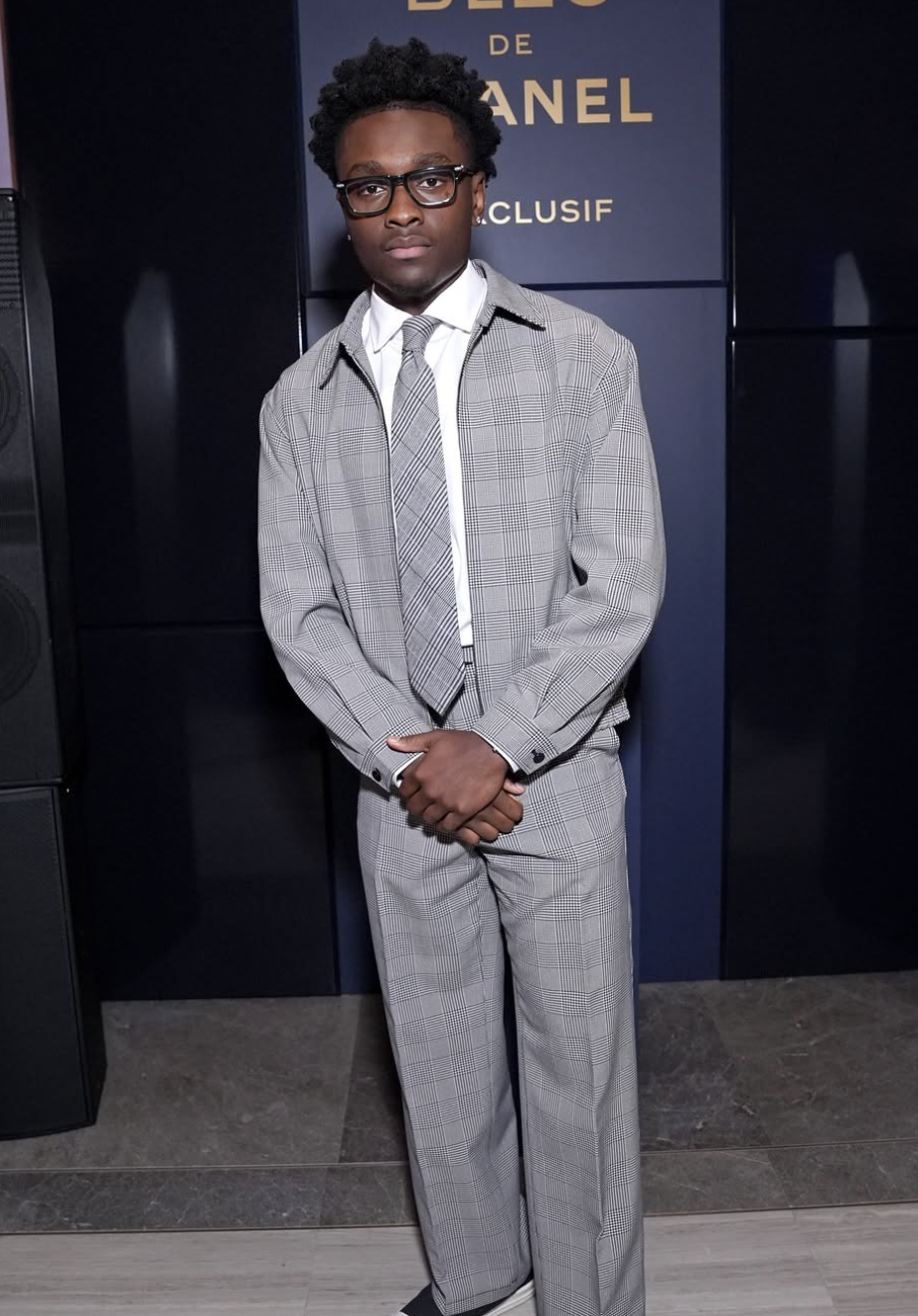 A man with curly hair, wearing glasses, a plaid suit with a white shirt and matching tie, standing indoors with a dark background and a sign that reads "DE CHANEL EXCLUSIF."