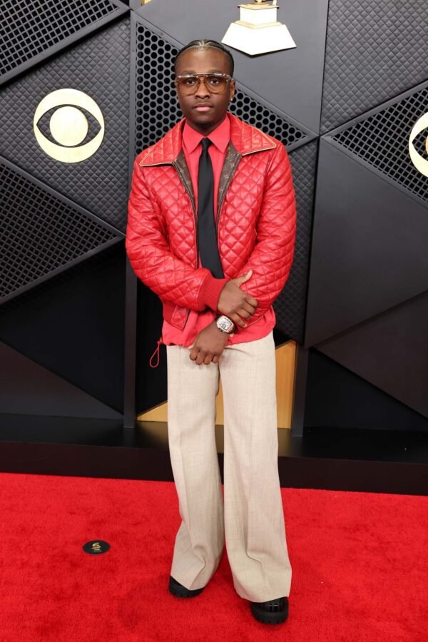 A man standing on a red carpet in front of a black backdrop with gold CBS logos. He is wearing a red quilted jacket, light-colored pants, a black shirt, and a watch, with sunglasses and a serious expression.