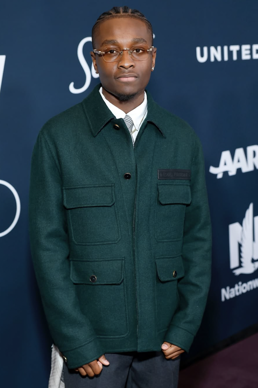 A young man with glasses, wearing a dark green jacket over a white shirt, standing against a blue backdrop with text and logos.