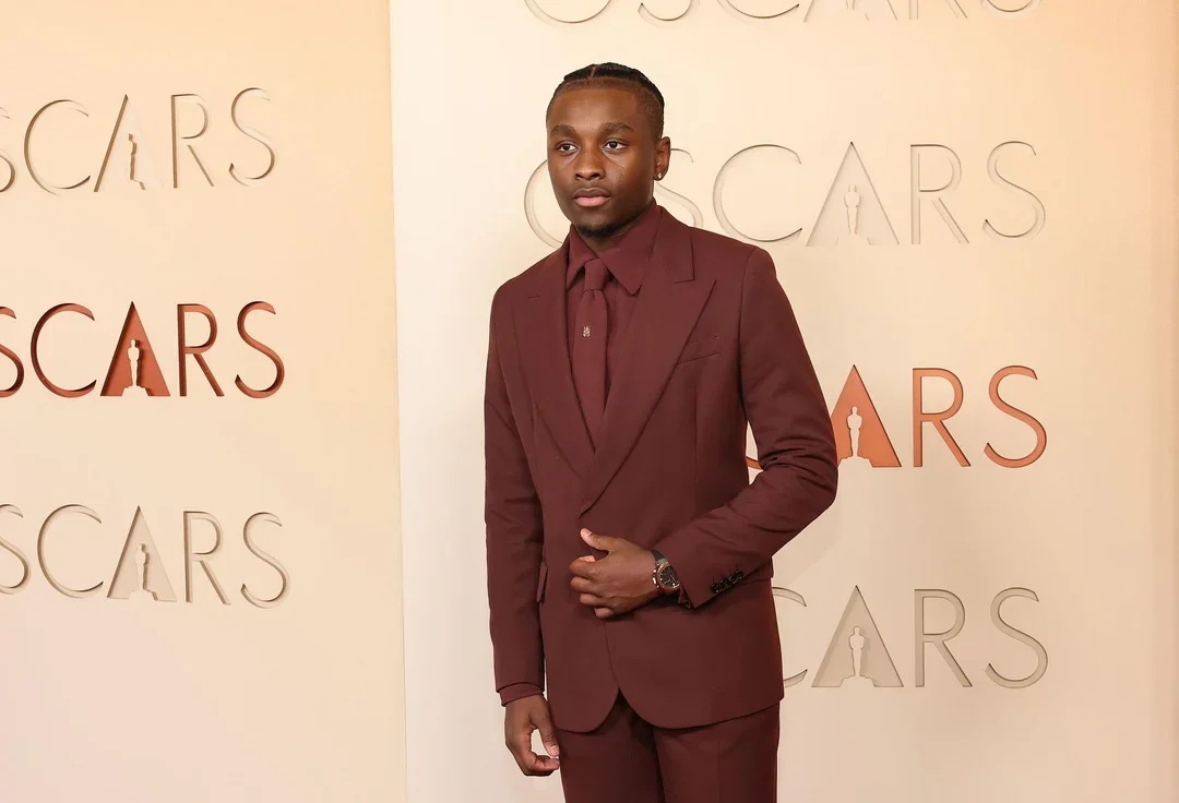 A young man in a maroon suit standing in front of an Oscars backdrop with repeated logos.