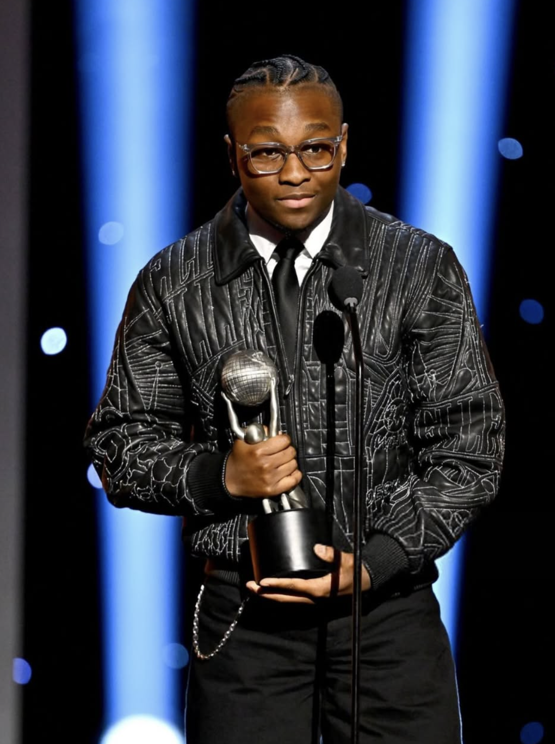 A young person in a black jacket holding an award statuette on stage at an awards ceremony.