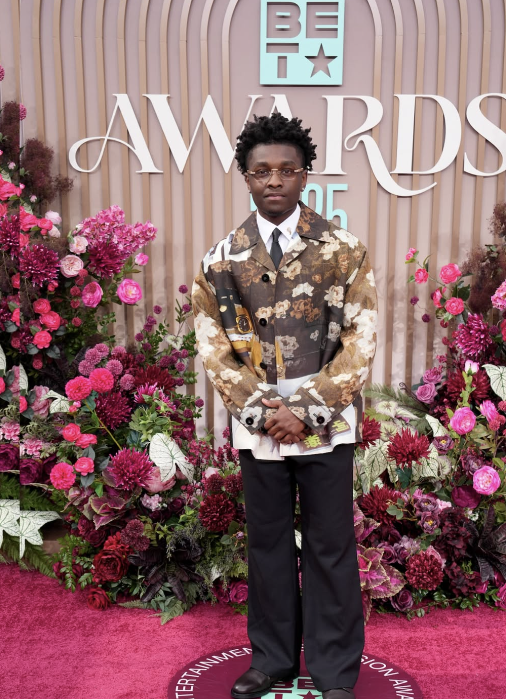 A man standing on a pink carpet at an awards event with floral arrangements behind him. He is wearing glasses, a patterned jacket, a white shirt, black pants, and black shoes.