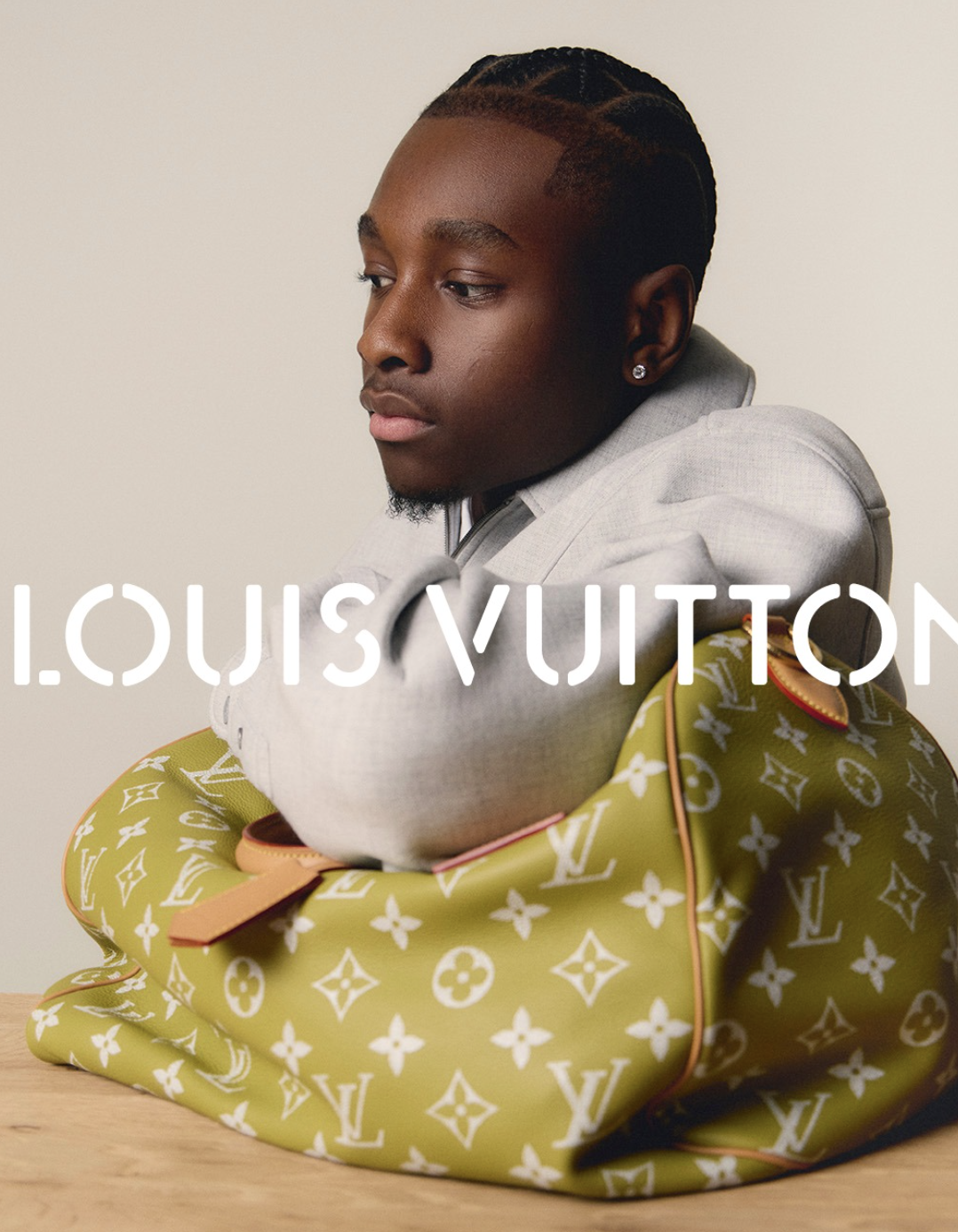 A young man with braided hair and earrings, wearing a light gray blazer, rests his chin on a Louis Vuitton bag with the brand’s monogram pattern, against a plain background.