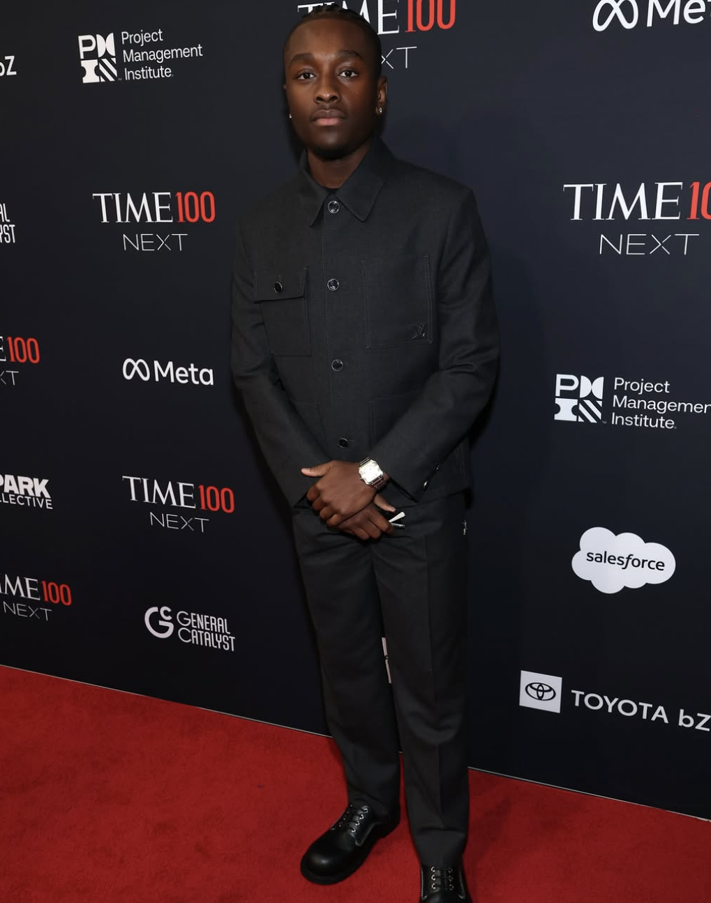 A young man in a black suit standing on a red carpet at a TIME 100 NEXT event, with logos of TIME, Meta, Project Management Institute, Salesforce, Toyota, and others visible on the backdrop.