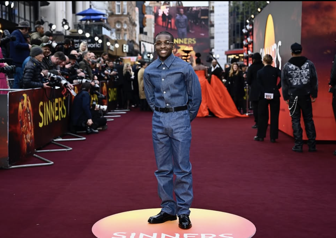 Young man standing on a red carpet at an event, smiling, wearing a denim outfit and surrounded by photographers and onlookers.