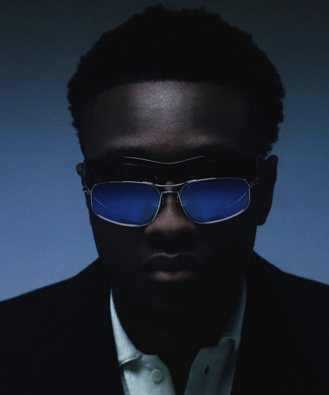 A person wearing sunglasses with blue-colored lenses, black suit, and white shirt, with a dark background