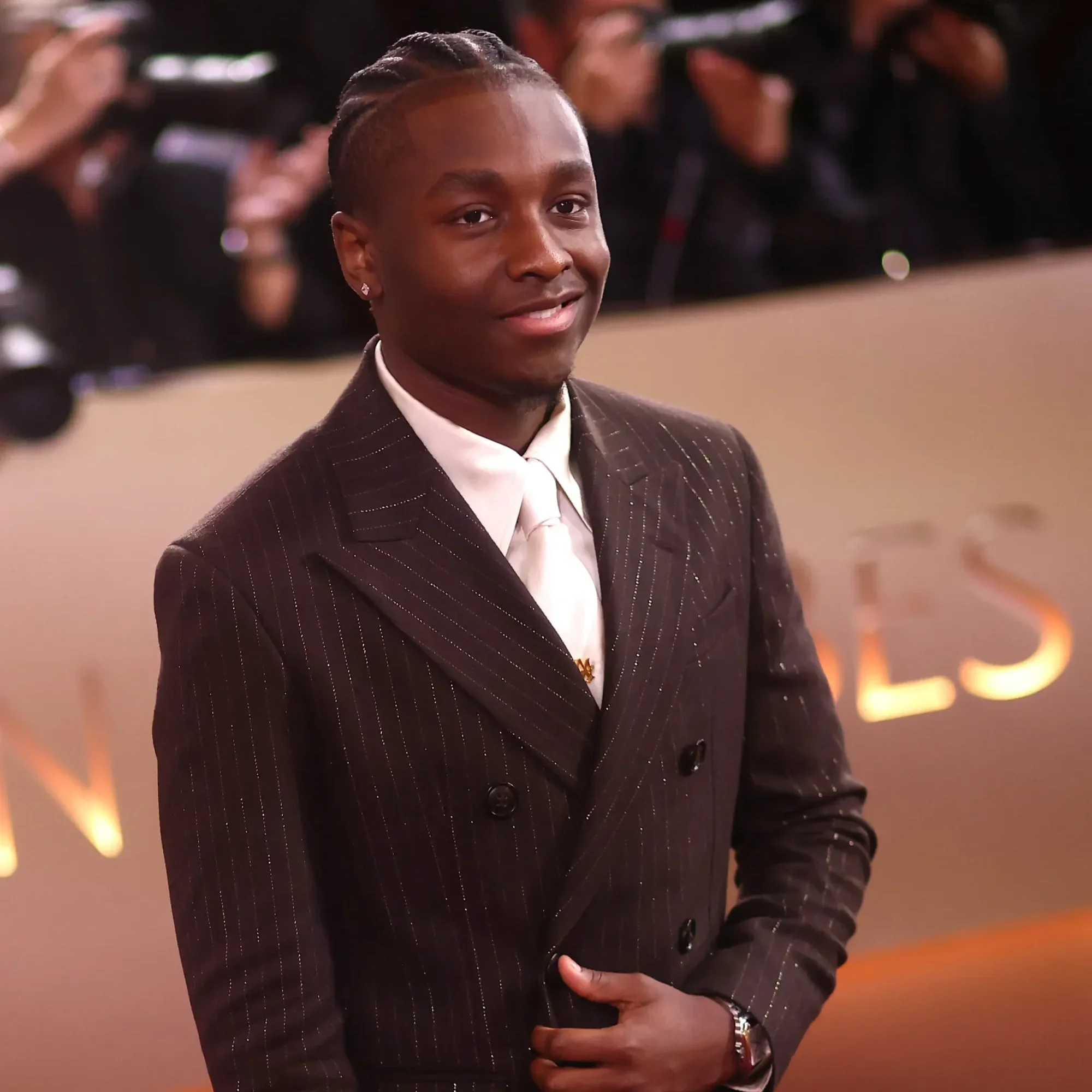 A smiling man dressed in a dark pinstripe suit, white shirt, and tie, with his arm around someone and friends in the background at a formal event.