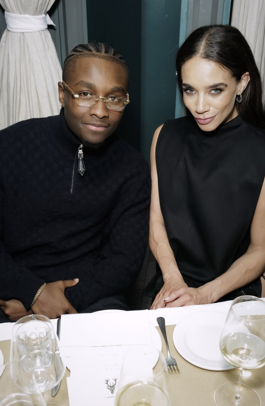 A man with glasses and braided hair wearing a black sweater, sitting next to a woman with dark hair, wearing a black sleeveless top, seated at a dining table with glassware and a menu.