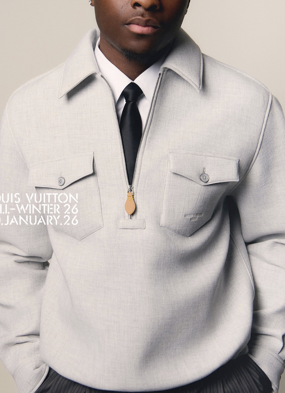 Close-up of a man wearing a light gray Louis Vuitton winter jacket, a white dress shirt, and a black tie, standing against a neutral background.