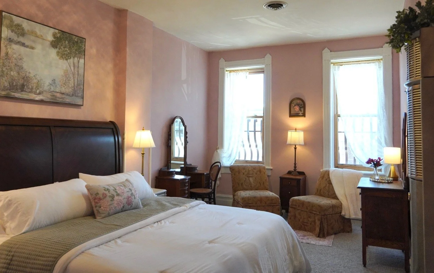 A cozy vintage-style bedroom with a large bed, wooden headboard, floral pillow, and cream-colored bedding. The room has pink walls, two large windows with sheer white curtains, and several lamps providing warm lighting. There are chairs, a small dresser, and a mirror against the wall.