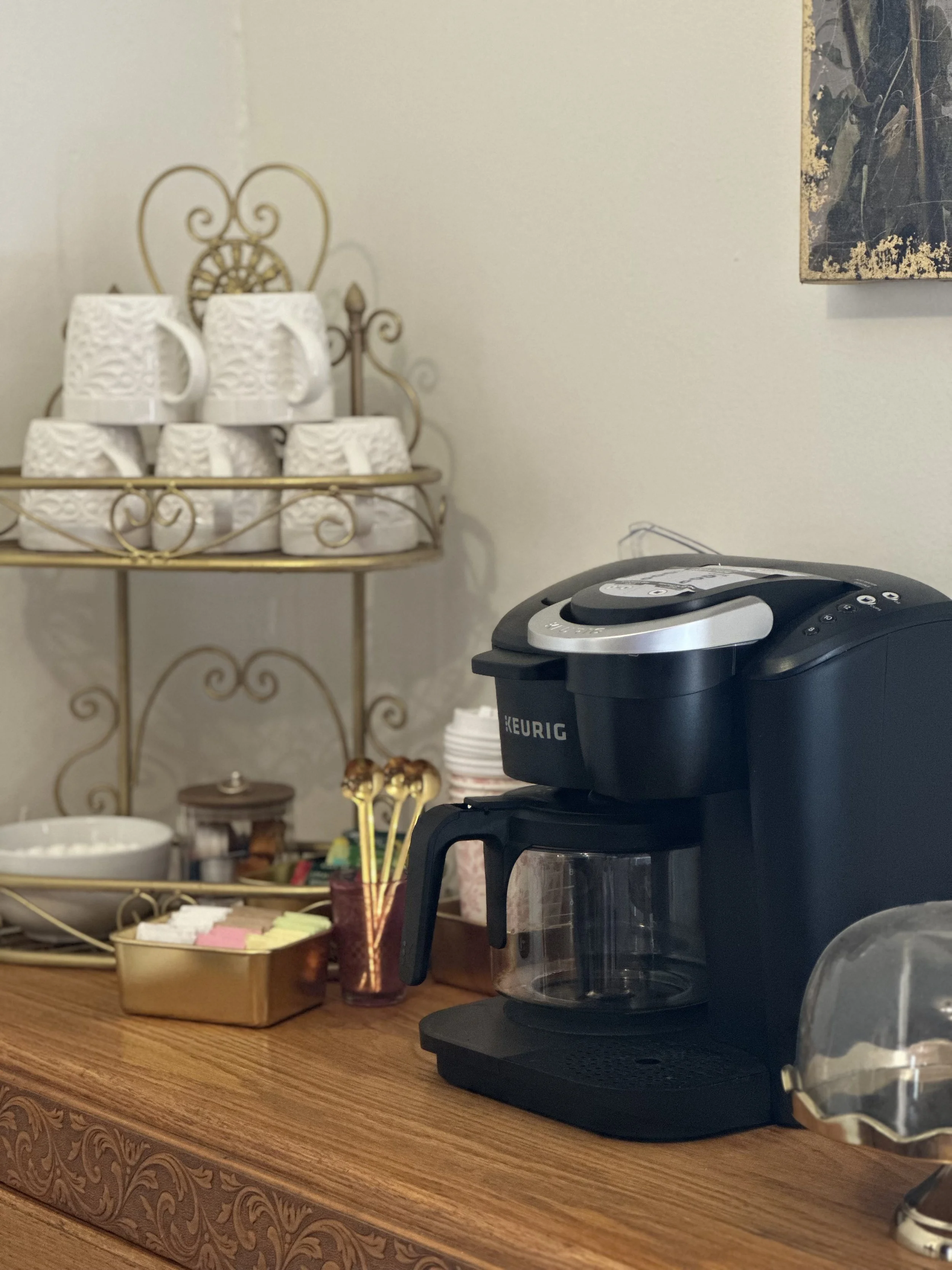 A kitchen countertop with a black coffee maker, a small tray of sugar packets, a decorative jar with lollipop sticks, a basket of sugar packets, and a wall-mounted metal shelf holding white coffee mugs, with a beige wall and a portion of a framed picture in the background.