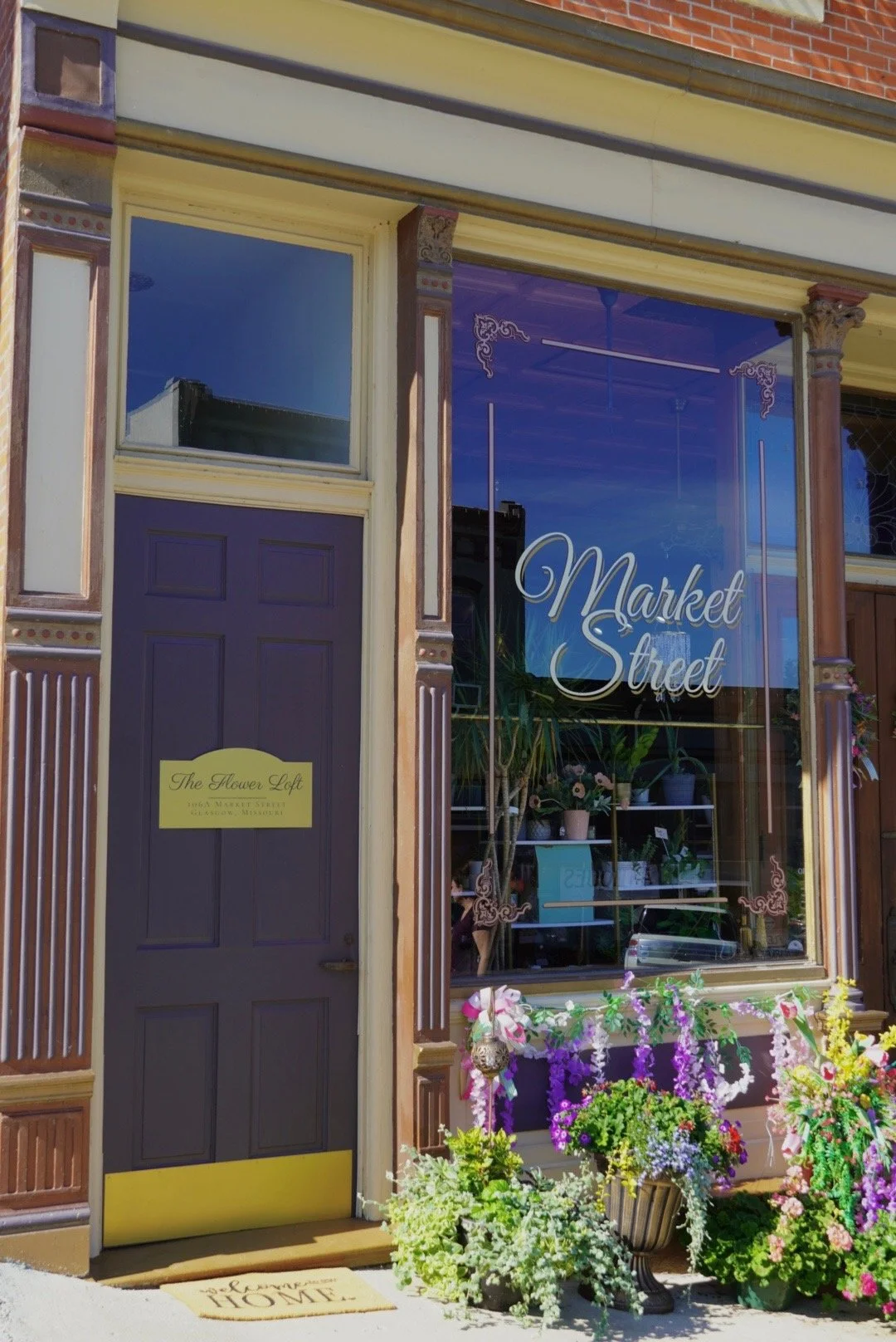 The storefront of 'Market Street' features a large window decorated with purple, pink, and white flowers in pots at the base. The door is painted dark purple with a yellow bottom panel, and has a yellow sign that reads 'The Flower Loft' with their address in Glasgow, Minnesota. A welcome mat with 'Welcome Home' written on it lies in front of the door. The building has ornate decorative trim and columns, and the street scene is reflected in the large glass window.