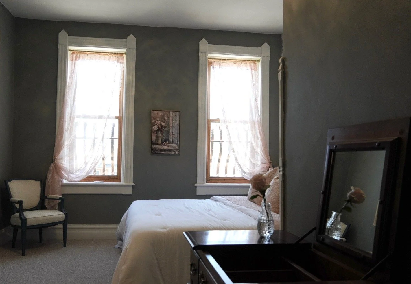 A bedroom with two windows that have sheer pink curtains. There is a bed with white bedding, a small chair with a dark frame and beige cushion, a framed picture on the wall, a dresser with a mirror, and a small vase with pink roses on the dresser.