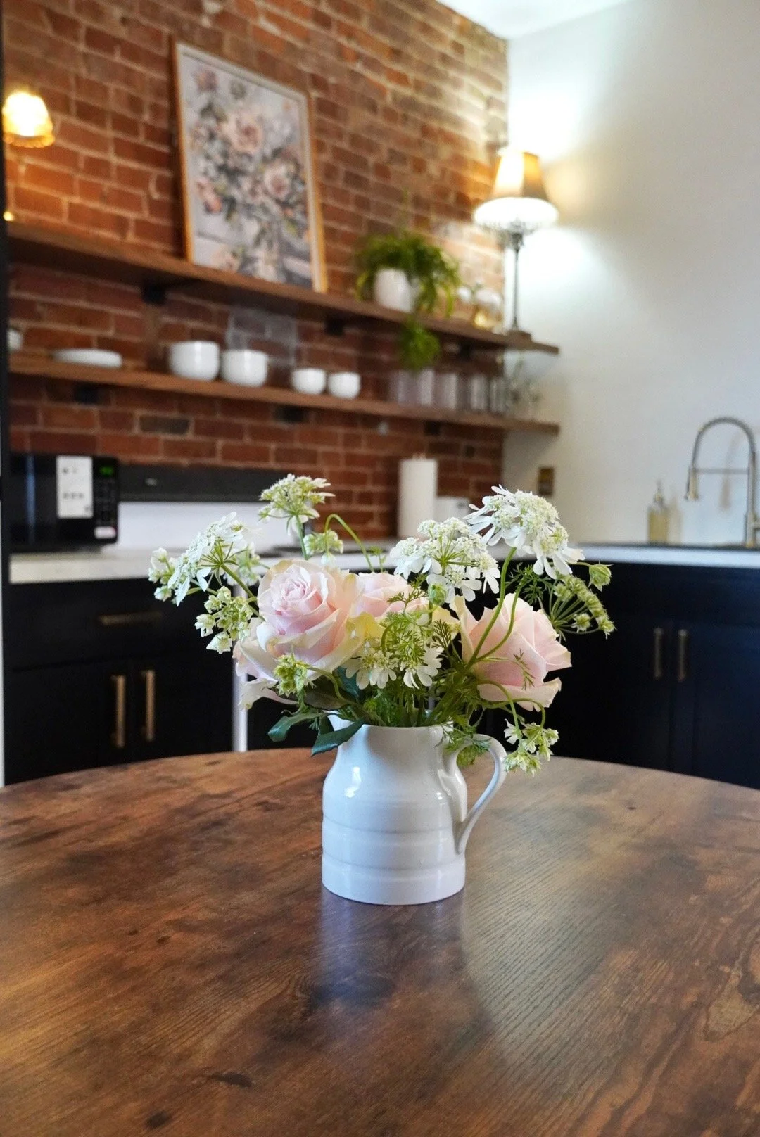 A white ceramic pitcher with light pink roses and small white flowers on a wooden table in a kitchen with exposed brick wall, shelves with dishes, and a stove in the background.