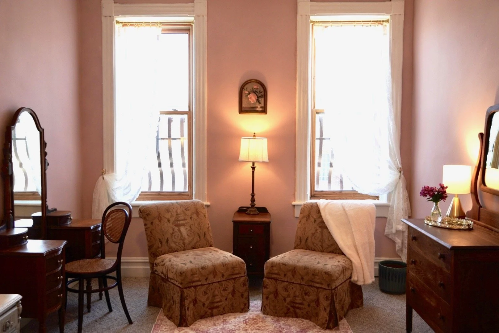 A cozy vintage-style living room with two large windows draped with sheer curtains, a small wooden side table with a table lamp, a pair of patterned armchairs, a wooden dresser with a lamp and a vase of flowers, and a decorative mirror on the wall.