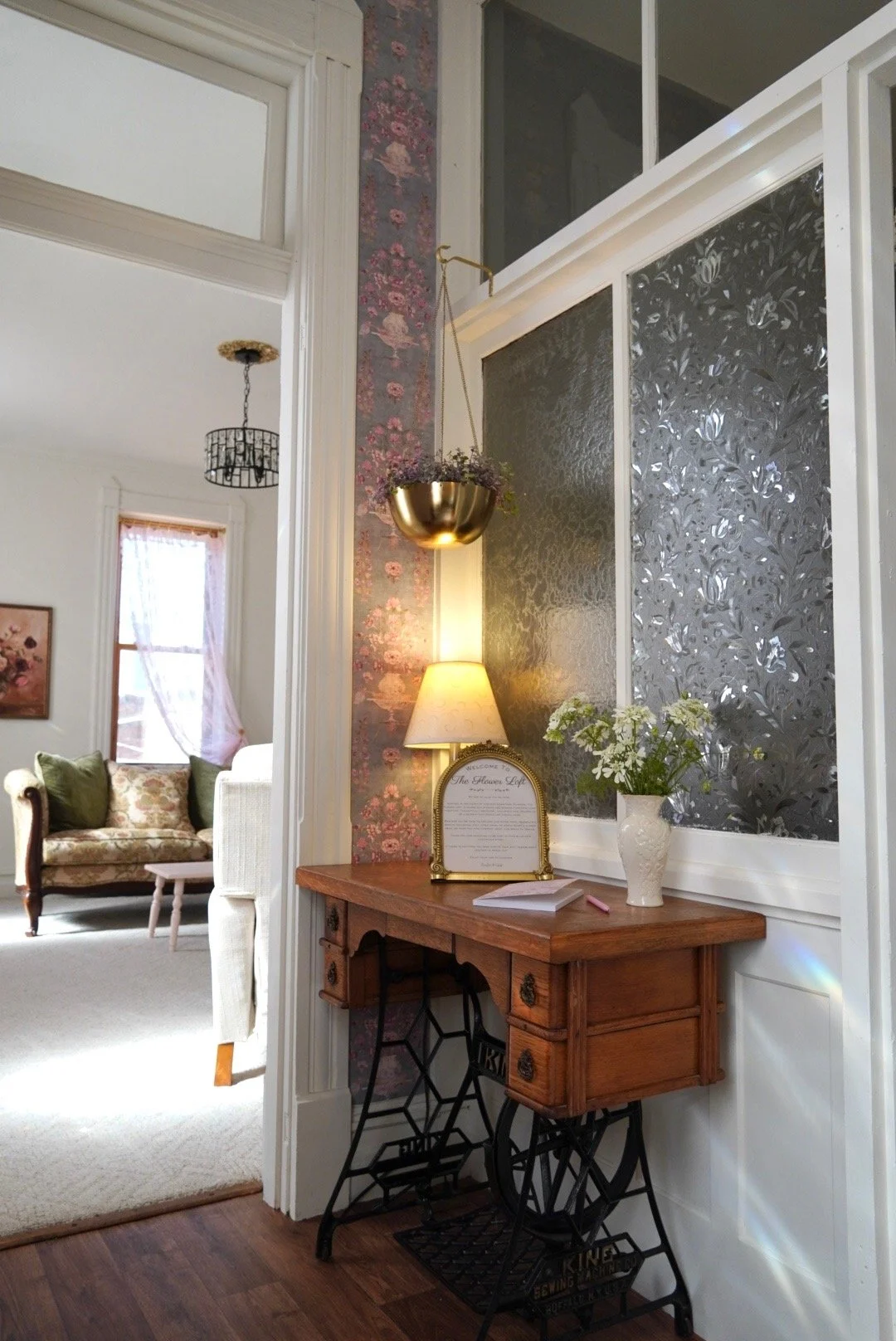 Interior view of a cozy living space with vintage decor, a sewing machine stand repurposed as a table with a wooden top, a potted white flower, a framed message, a lamp, and decorative wall panels, with a sitting area in the background.