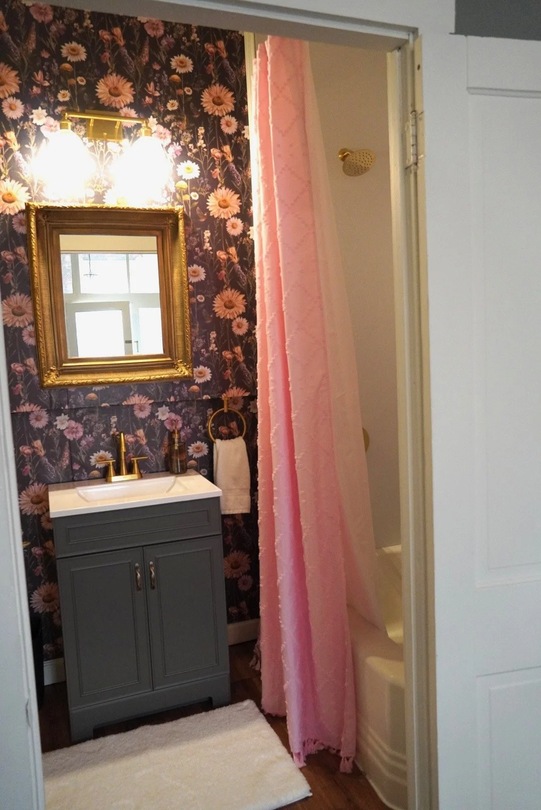 A small bathroom with floral wallpaper, a gray vanity with a white sink, gold fixtures, and a gold-framed mirror. There is a pink shower curtain and a pink shower wall inside the shower area. A white towel hangs on a gold ring beside the vanity.