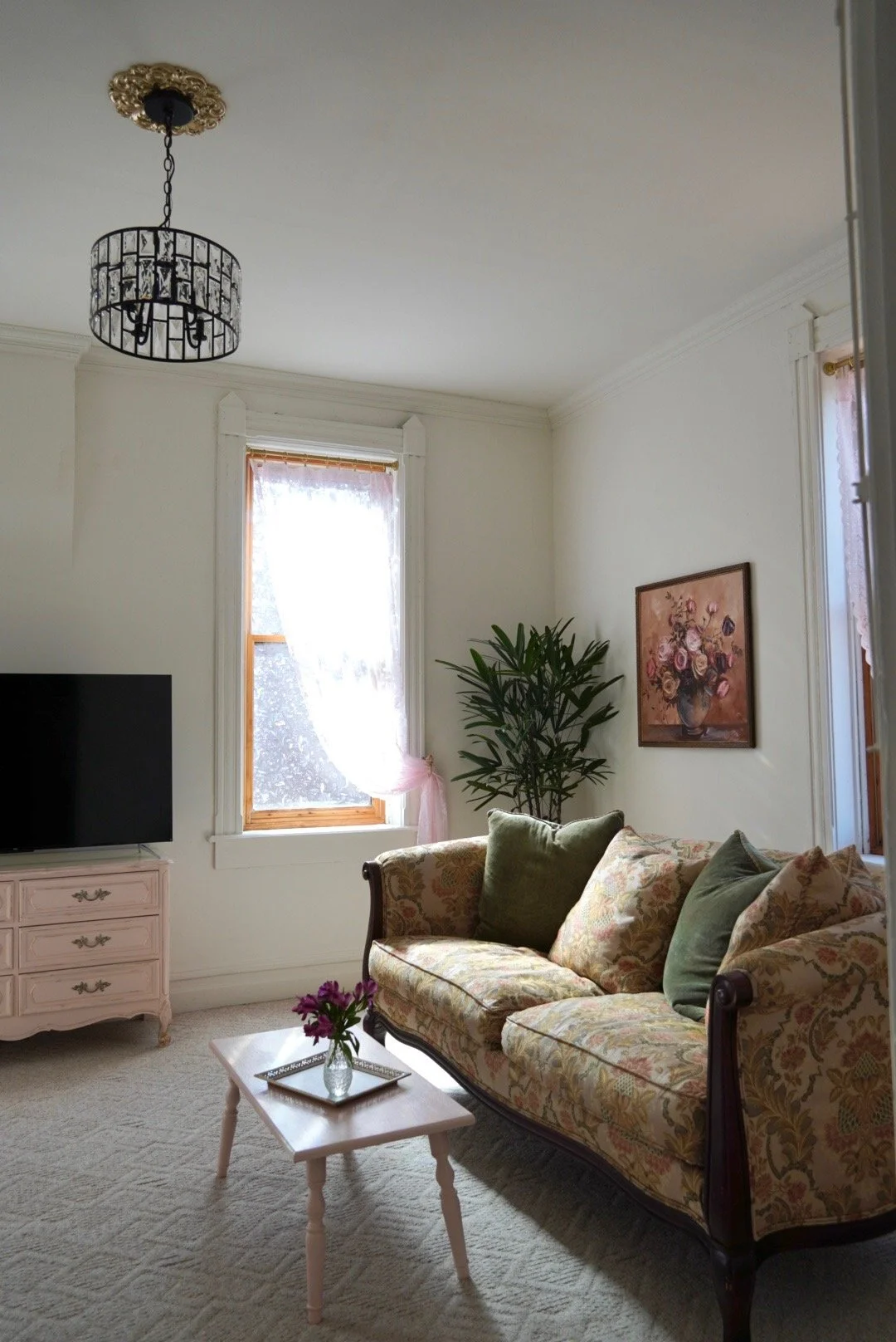 Living room with a floral sofa, green pillows, a small white table with a flower vase, a pink dresser, a flat-screen TV, and a framed floral painting on the wall. Two windows with pink curtains and a potted plant near the windows.